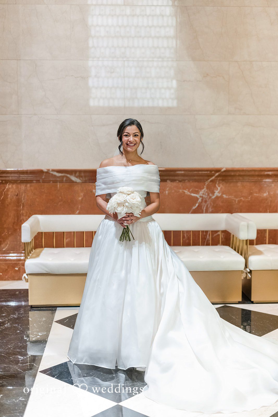 A bride in a white off-the-shoulder wedding gown smiling and holding a bouquet.