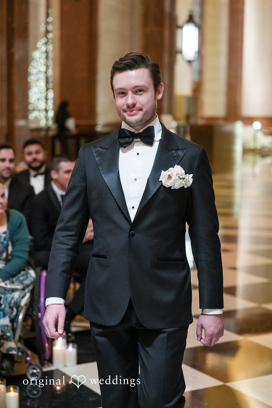 A groom walking down the aisle wearing a classic black tuxedo and bow tie.