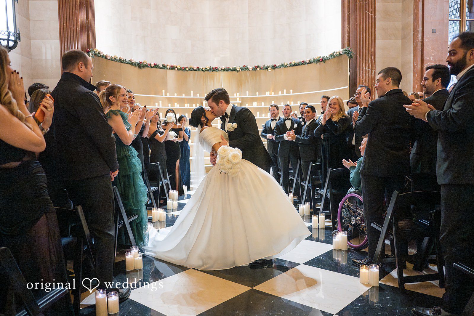 A bride and groom sharing a kiss at the end of the aisle while guests applaud.