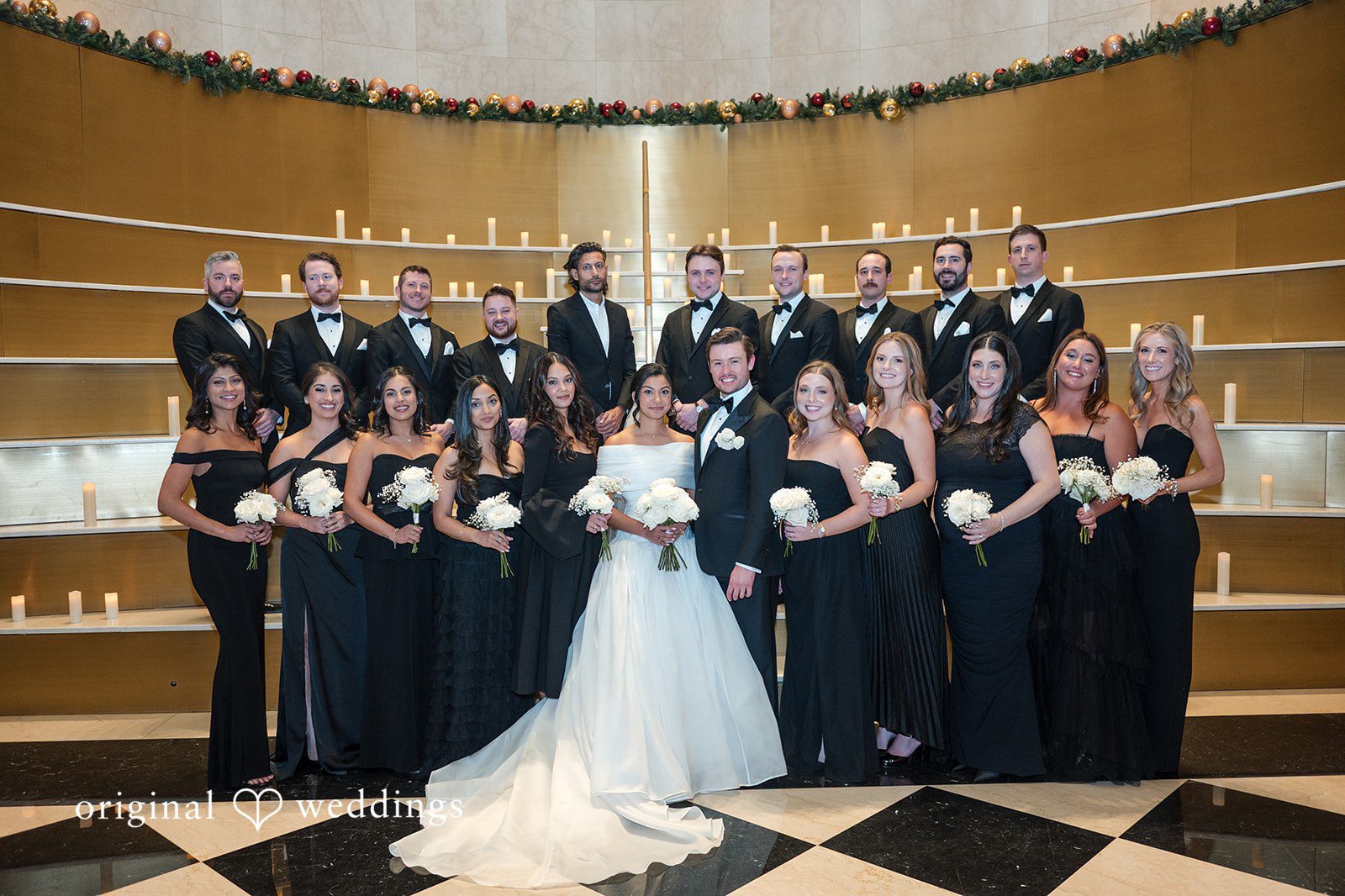 A large wedding party in black attire posing together on a checkered floor during a reception.