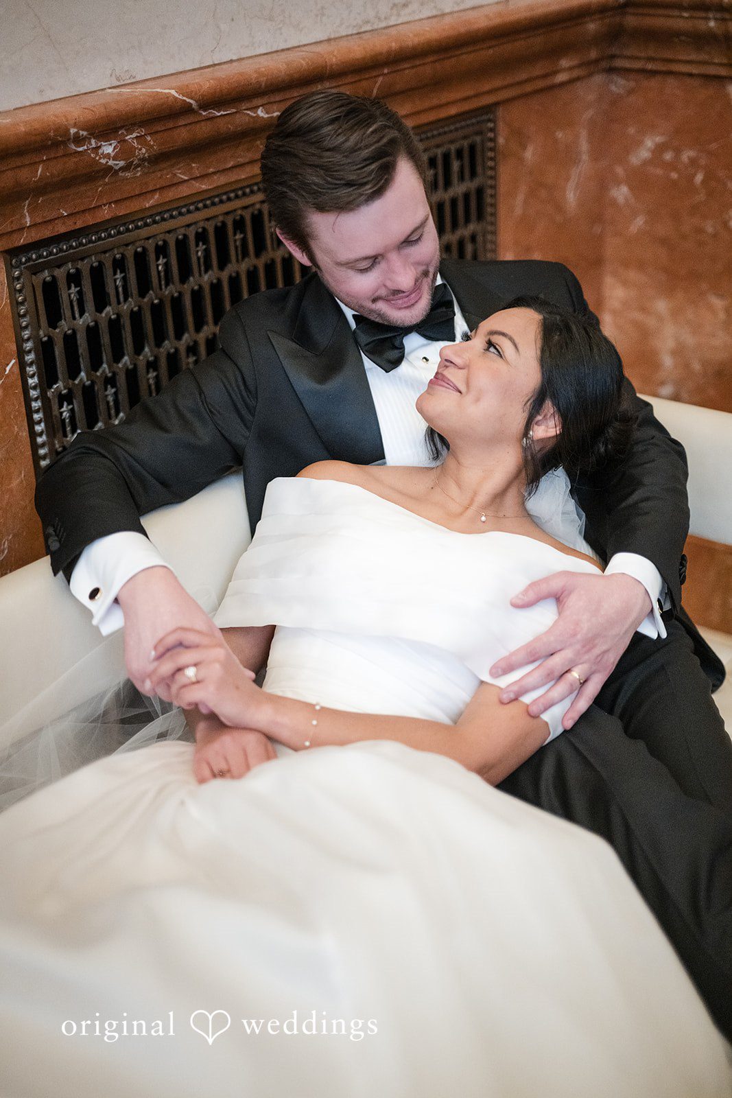 A newlywed couple sitting closely together, with the groom wrapping his arms around the bride.