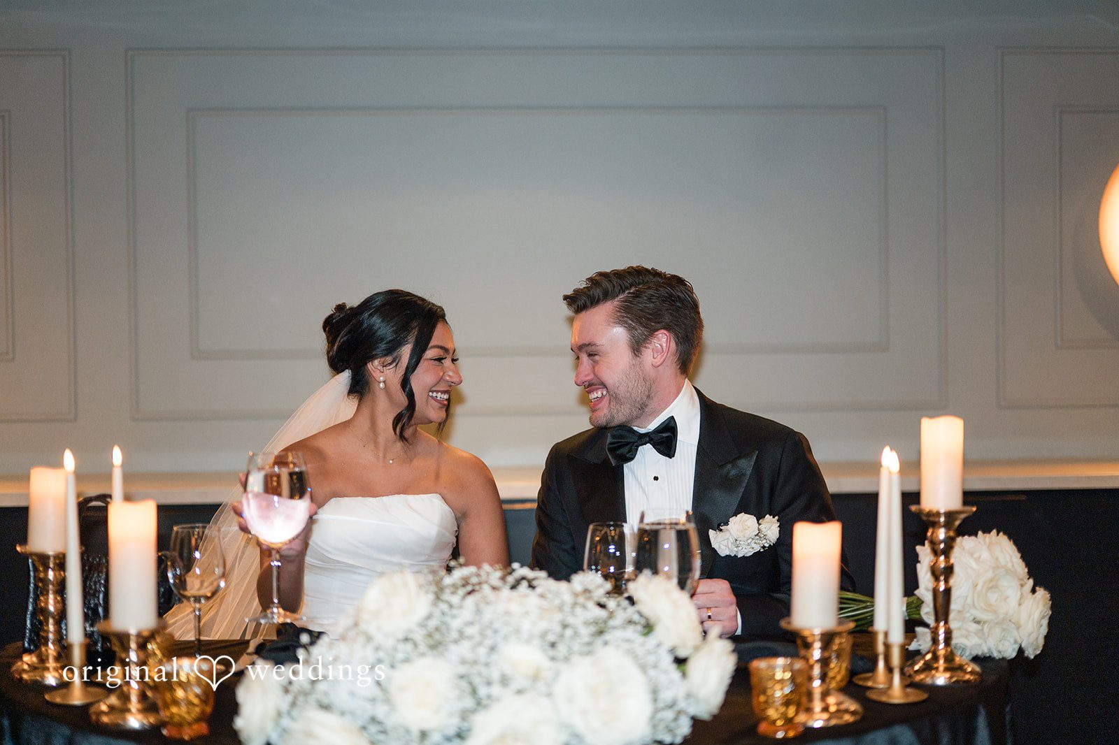 bride and groom sitting at their reception table decorated with white flowers and lit candles.