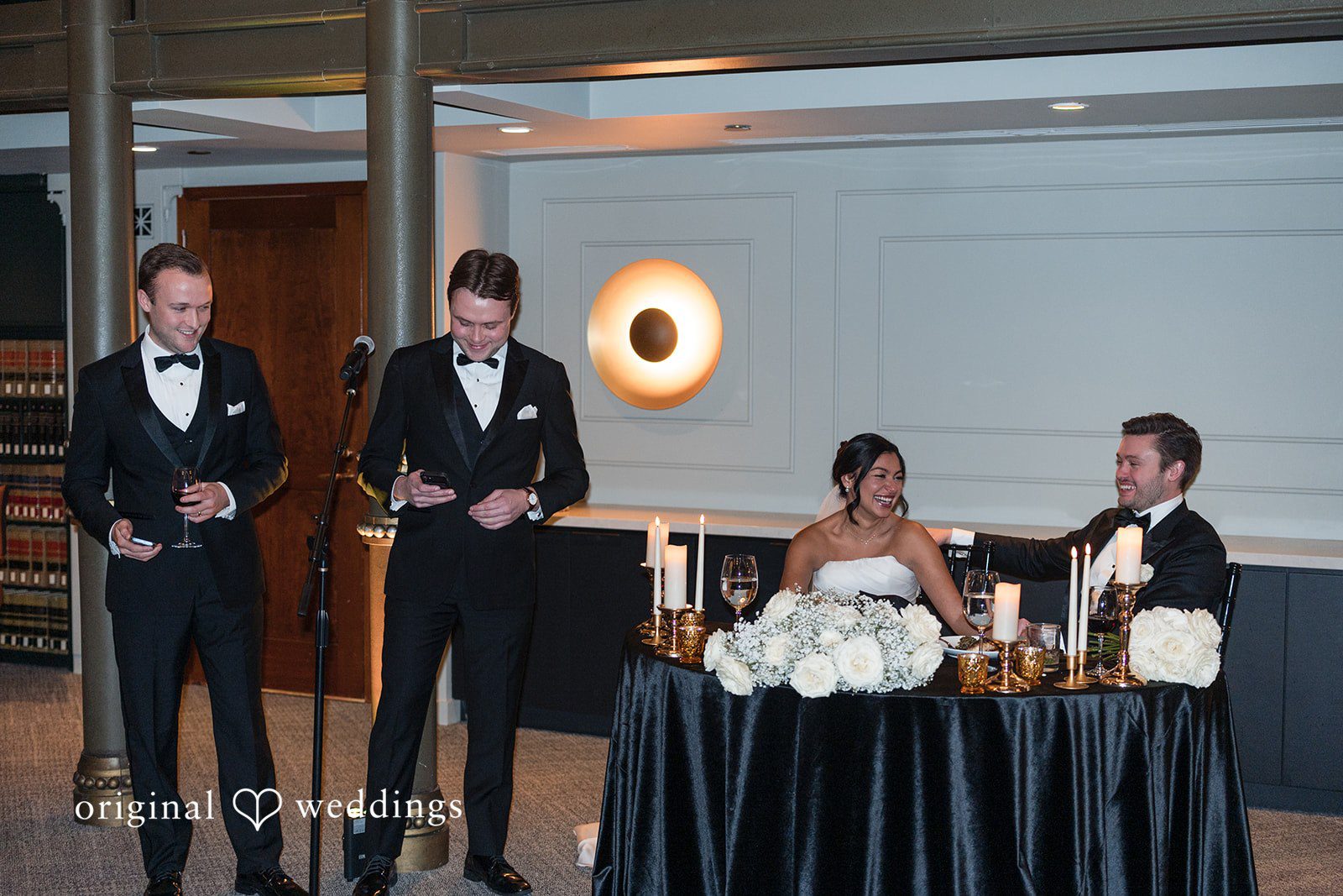 A bride and groom listening to a wedding speech being delivered by two men at the reception.