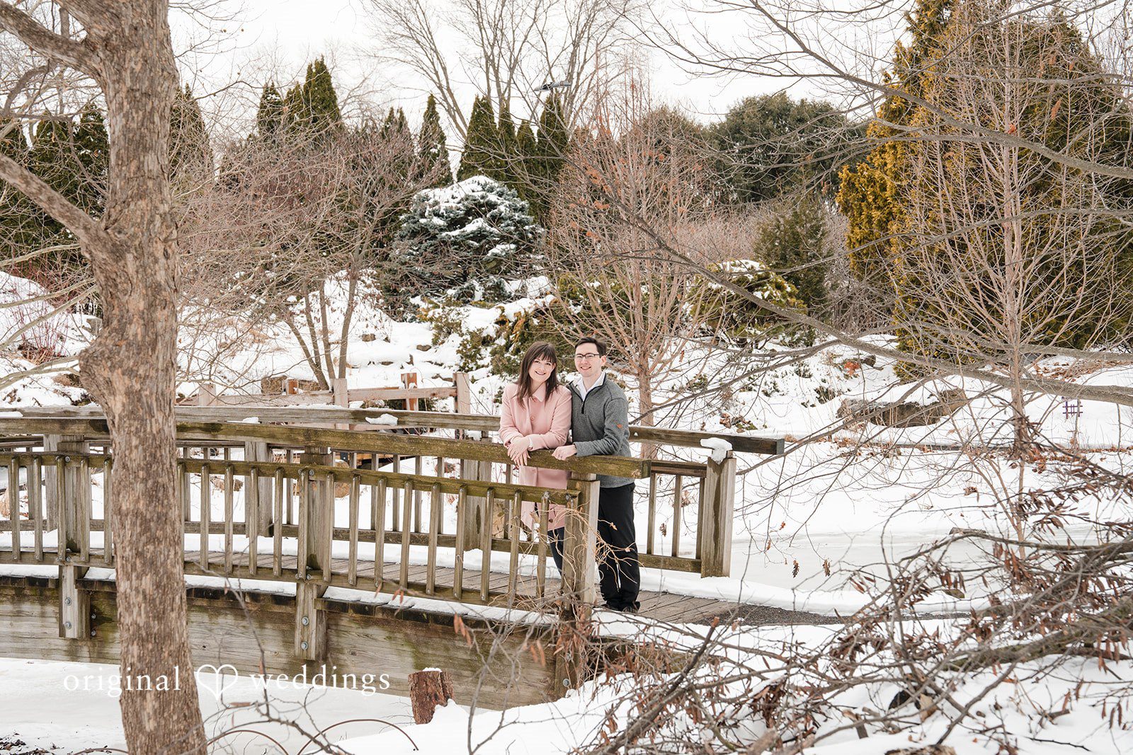 Engagement photography services by Original Weddings, capturing the couple standing together on a wooden bridge at Olbrich Gardens