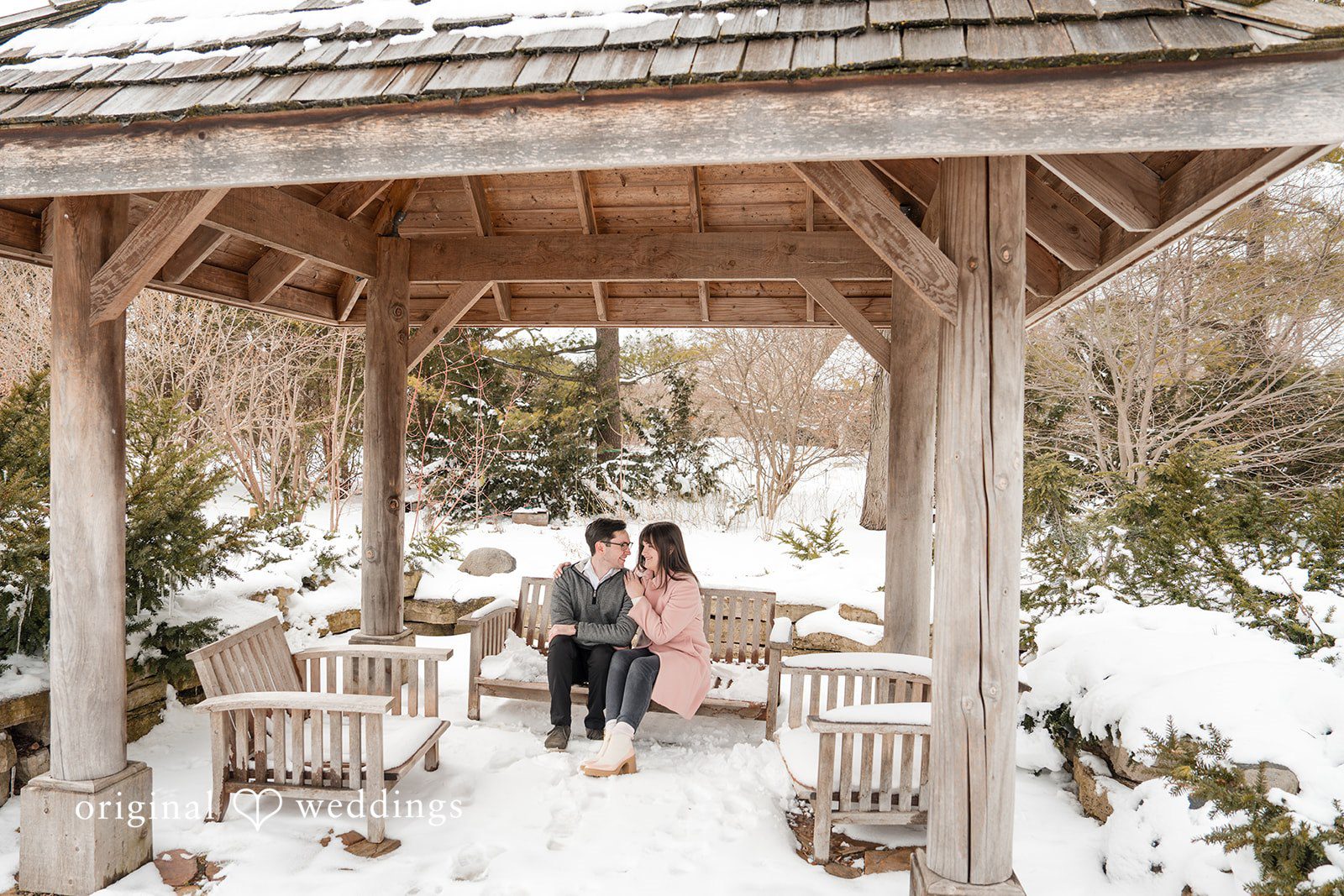 Engagement photography services by Original Weddings, capturing the couple sharing a playful moment under a gazebo at Olbrich Gardens, surrounded by snow-covered benches and trees