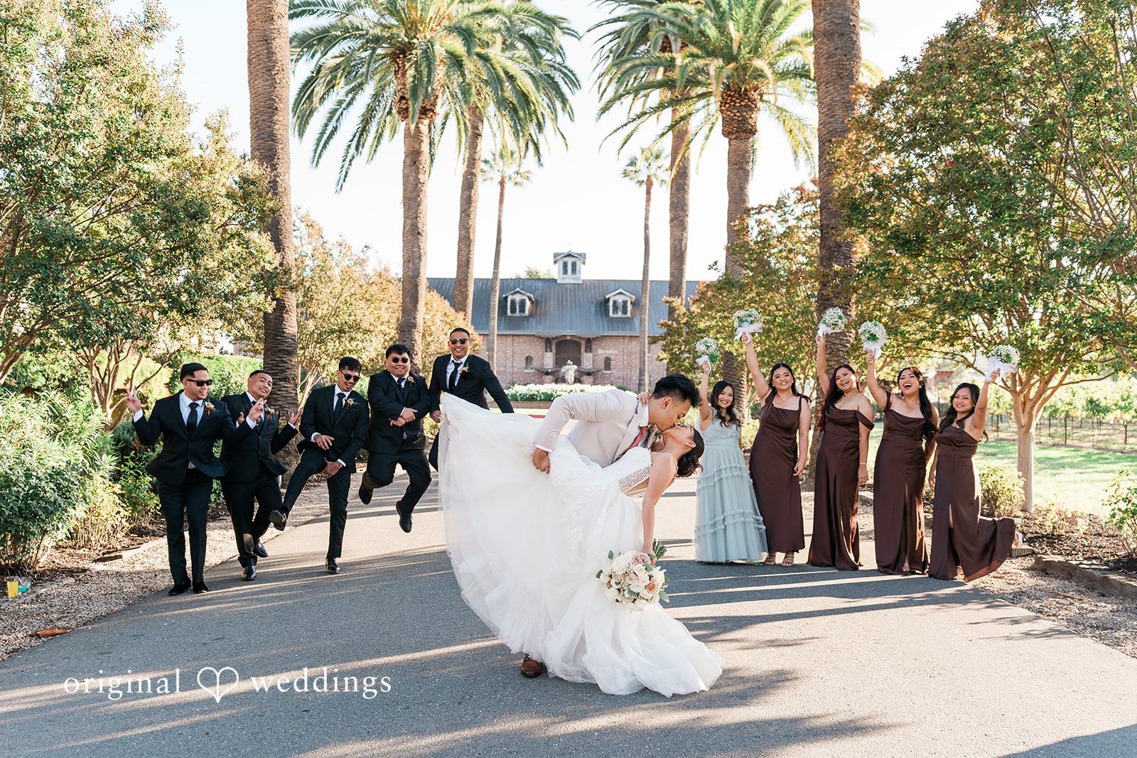 Charize Giann + Mark Shou Bride and groom sharing a romantic dip kiss with their wedding party cheering behind them at Palm Event Center captured by San Francisco wedding photographers from Original Weddings