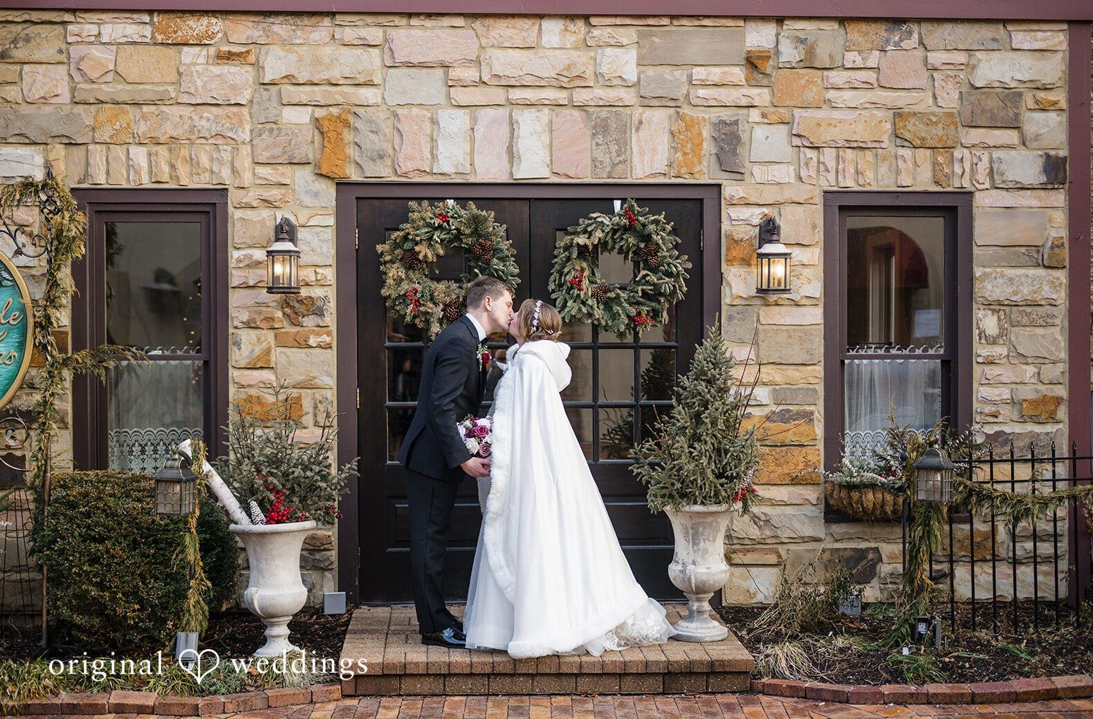 Chicago Wedding Photographers capture a timeless winter couple portrait at Riverside Receptions entrance with stone walls, wreath decor, and elegant formal attire.