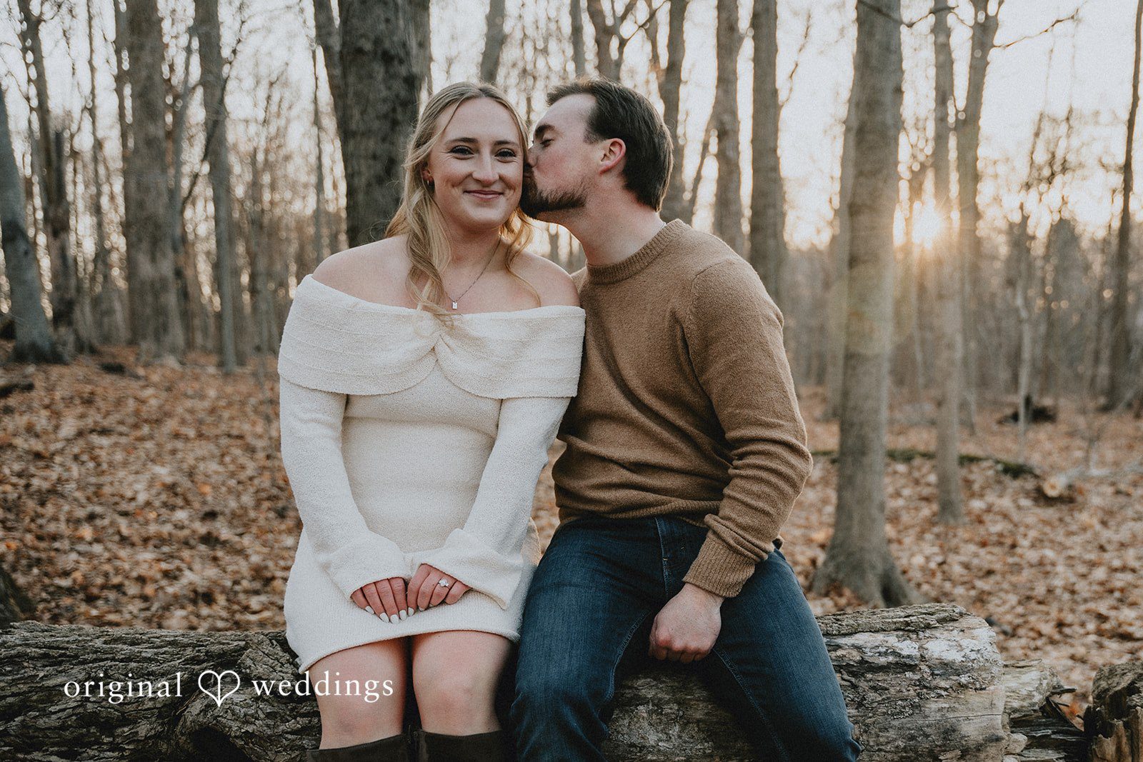 The groom kisses the bride's cheek while sitting on a log in a forest.