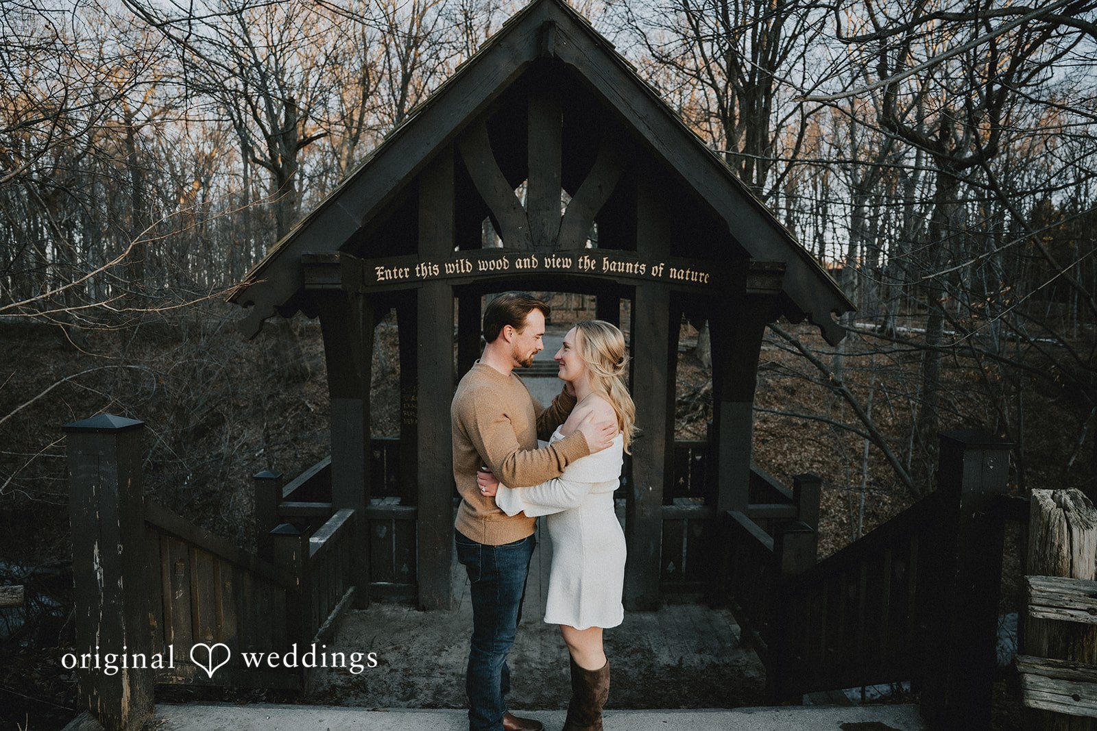 The couple sharing a romantic embrace in front of a rustic wooden gazebo in the woods.