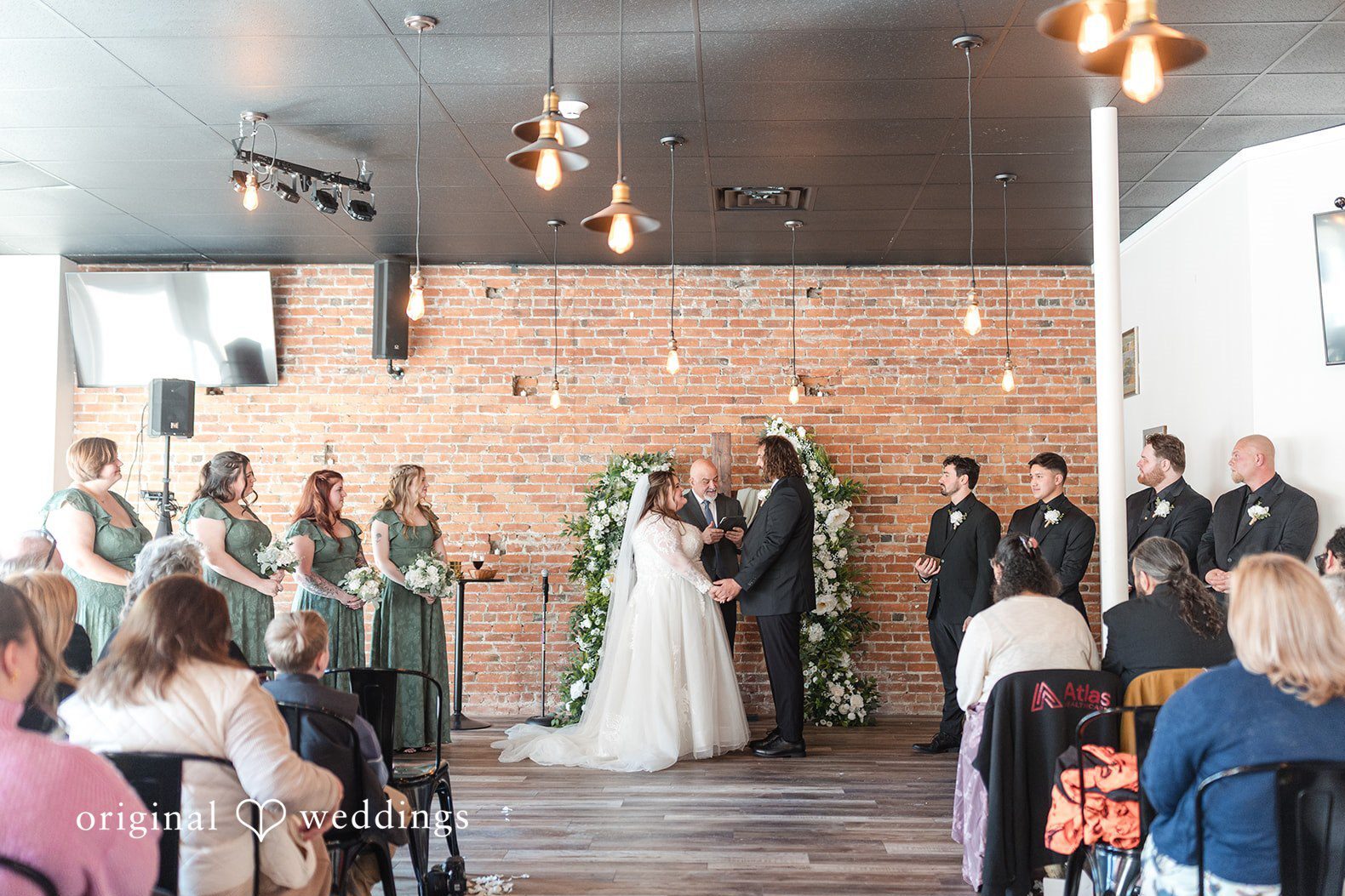 Indoor ceremony scene at The Bridge Church Middleboro, captured with Boston wedding photography, featuring a floral arch, brick walls, and guests gathered around the couple.