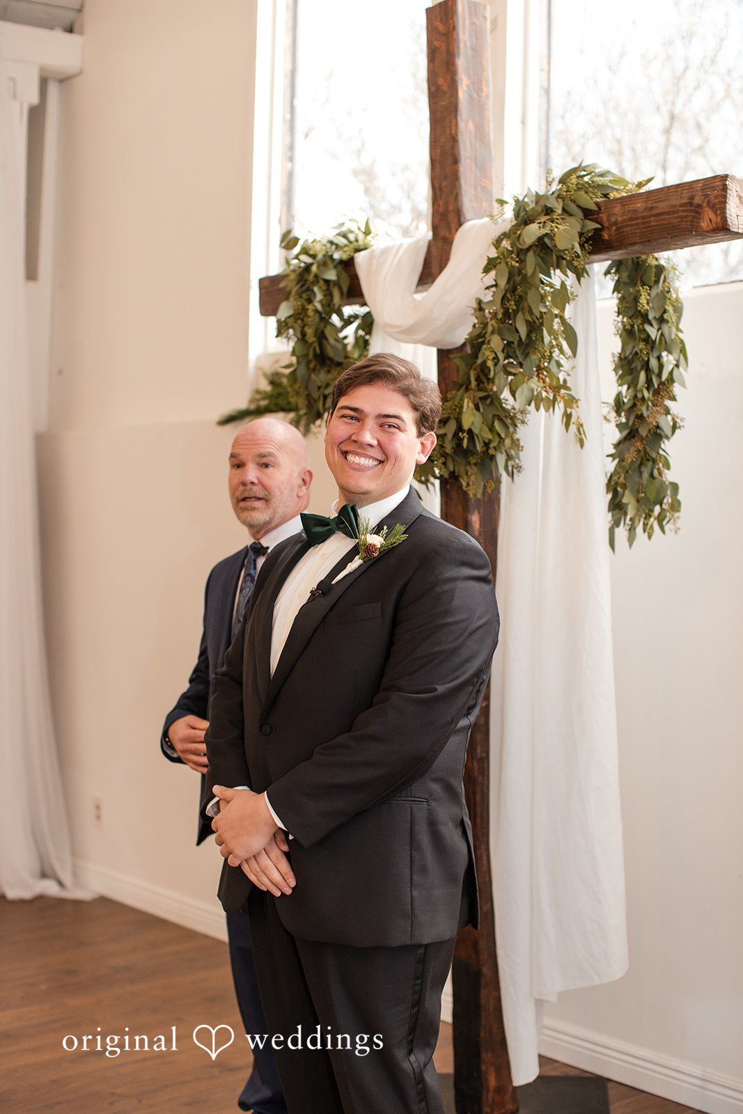 Adam + McKenzie Groom standing under ceremony arch decorated with greenery