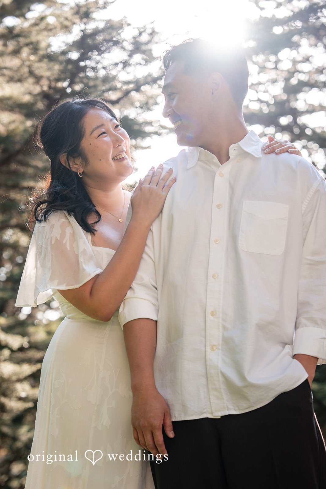 Engaged couple standing close, holding hands, and smiling in a forest setting