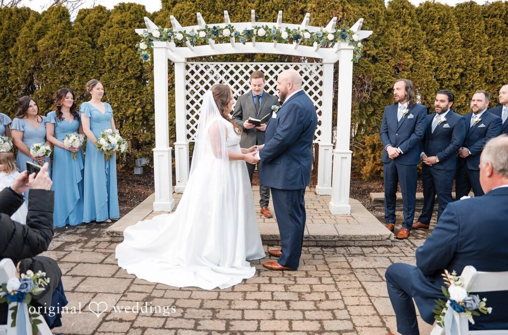 Detroit wedding photographers capturing an outdoor wedding ceremony under a white arbor at Zuccaro’s with blue themed bridal party and elegant setup