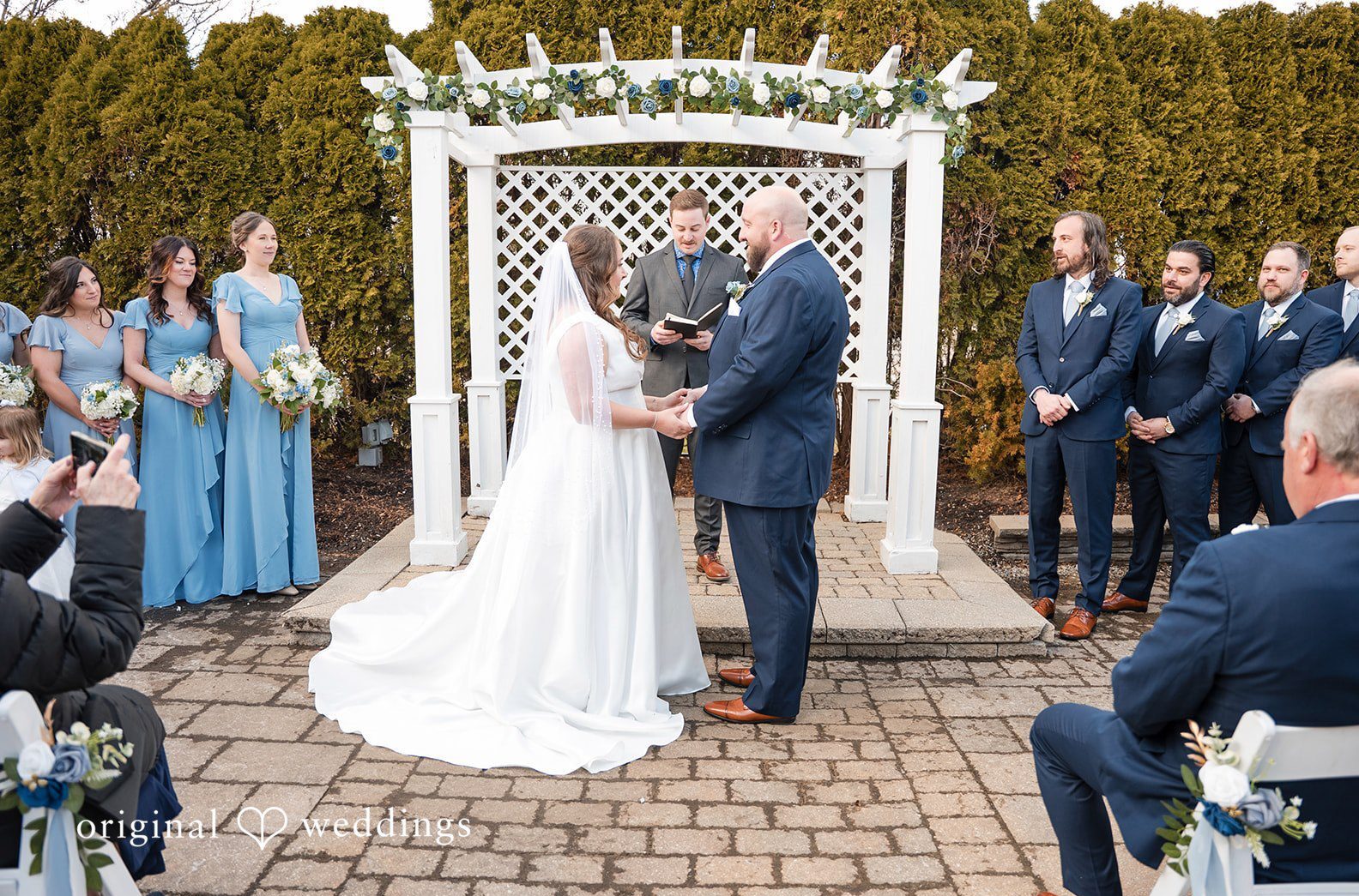 Detroit wedding photographers capturing an outdoor wedding ceremony under a white arbor at Zuccaro’s with blue themed bridal party and elegant setup