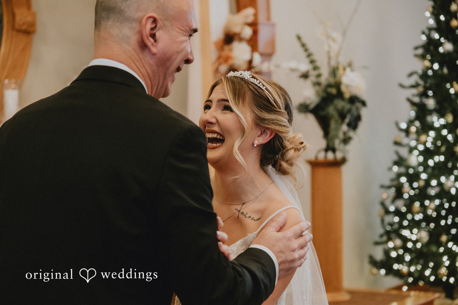 Sarah + Justin A bride laughs joyfully while looking up at a man in a black suit with a lit Christmas tree in the background.