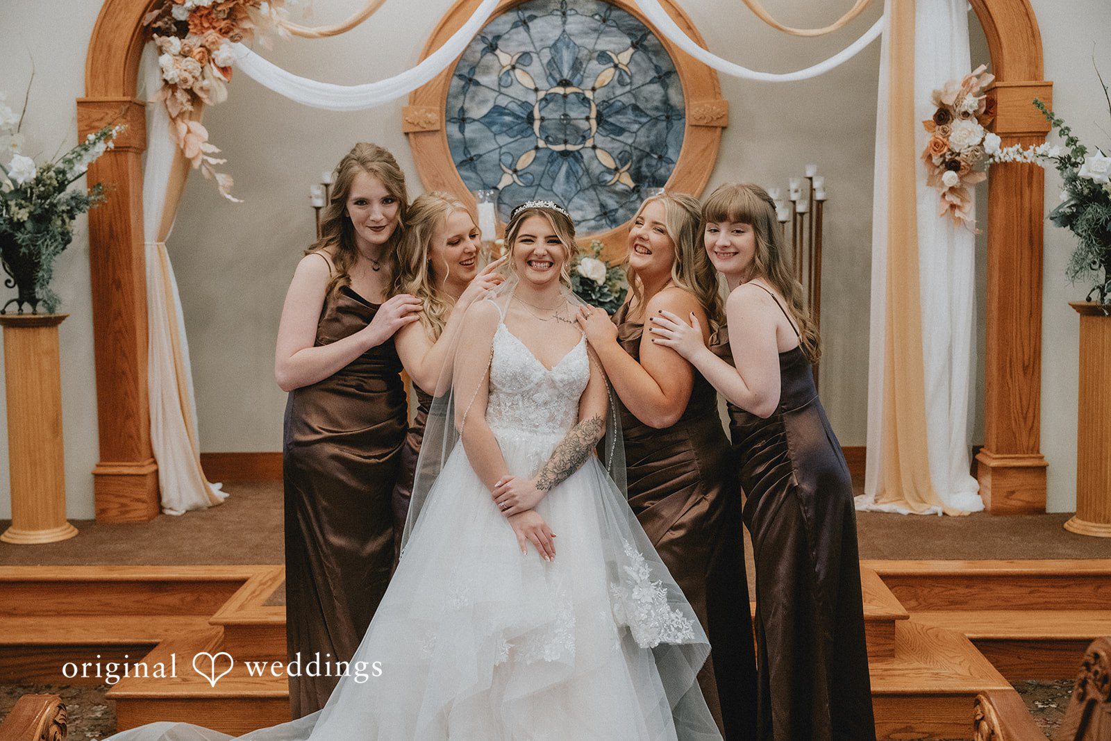 Sarah + Justin A smiling bride in a white gown and veil poses with four bridesmaids in brown satin dresses at an altar.