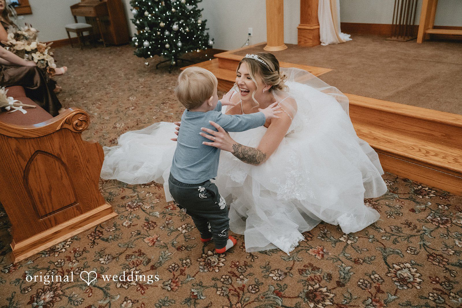 Sarah + Justin A laughing bride in a white gown crouches down to embrace a toddler in a church.