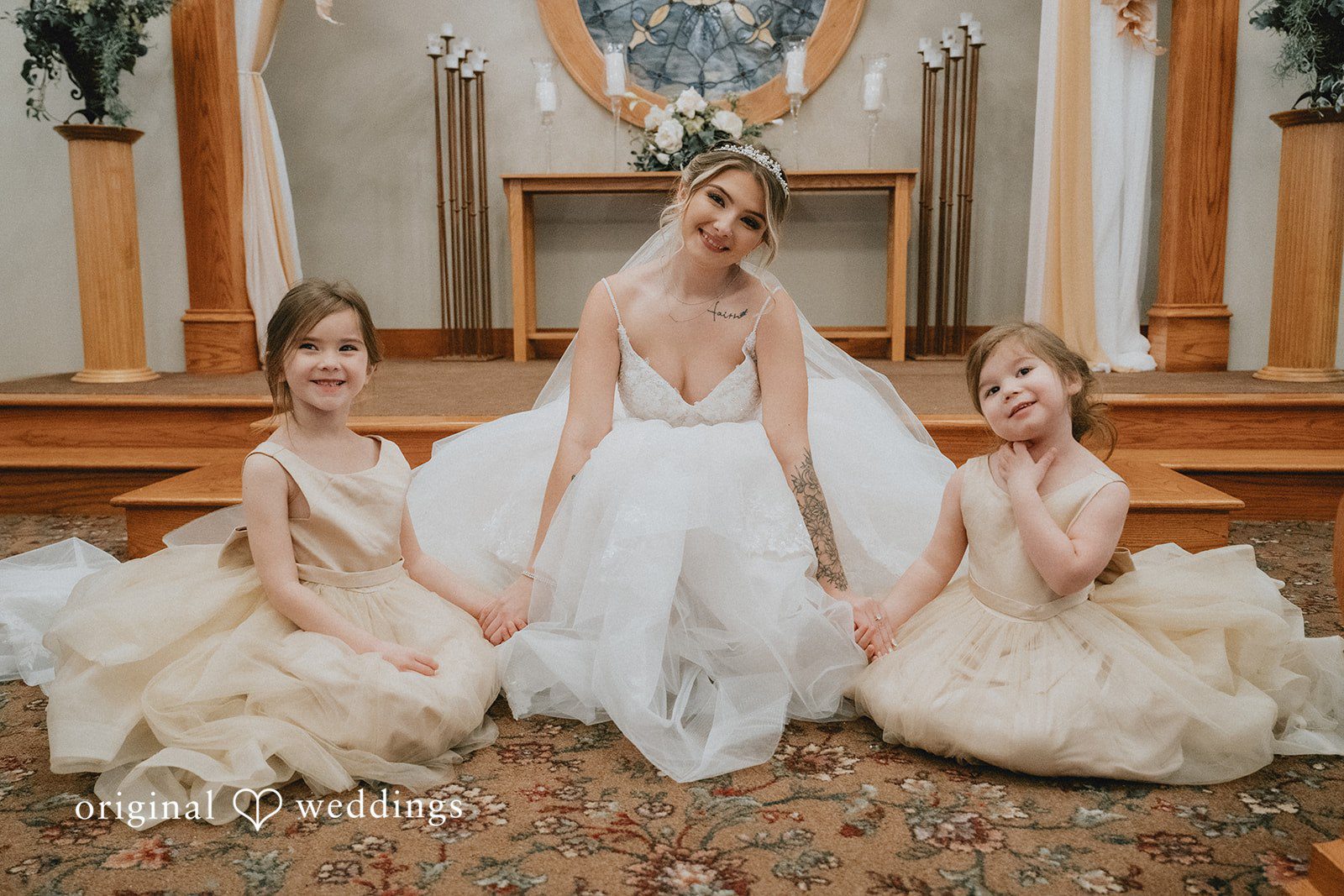 Sarah + Justin A smiling bride sits on the floor holding hands with two young girls in beige tulle dresses.