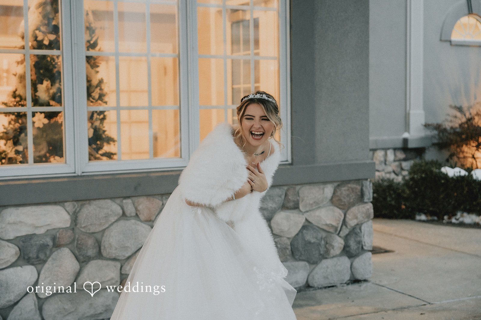 Sarah + Justin A laughing bride wearing a white gown, a tiara, and a white faux fur shawl stands outside a building.