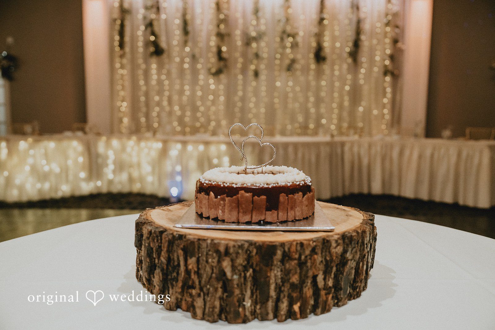 Sarah + Justin Rustic wedding cake displayed on a wooden stand with shimmering backdrop details.