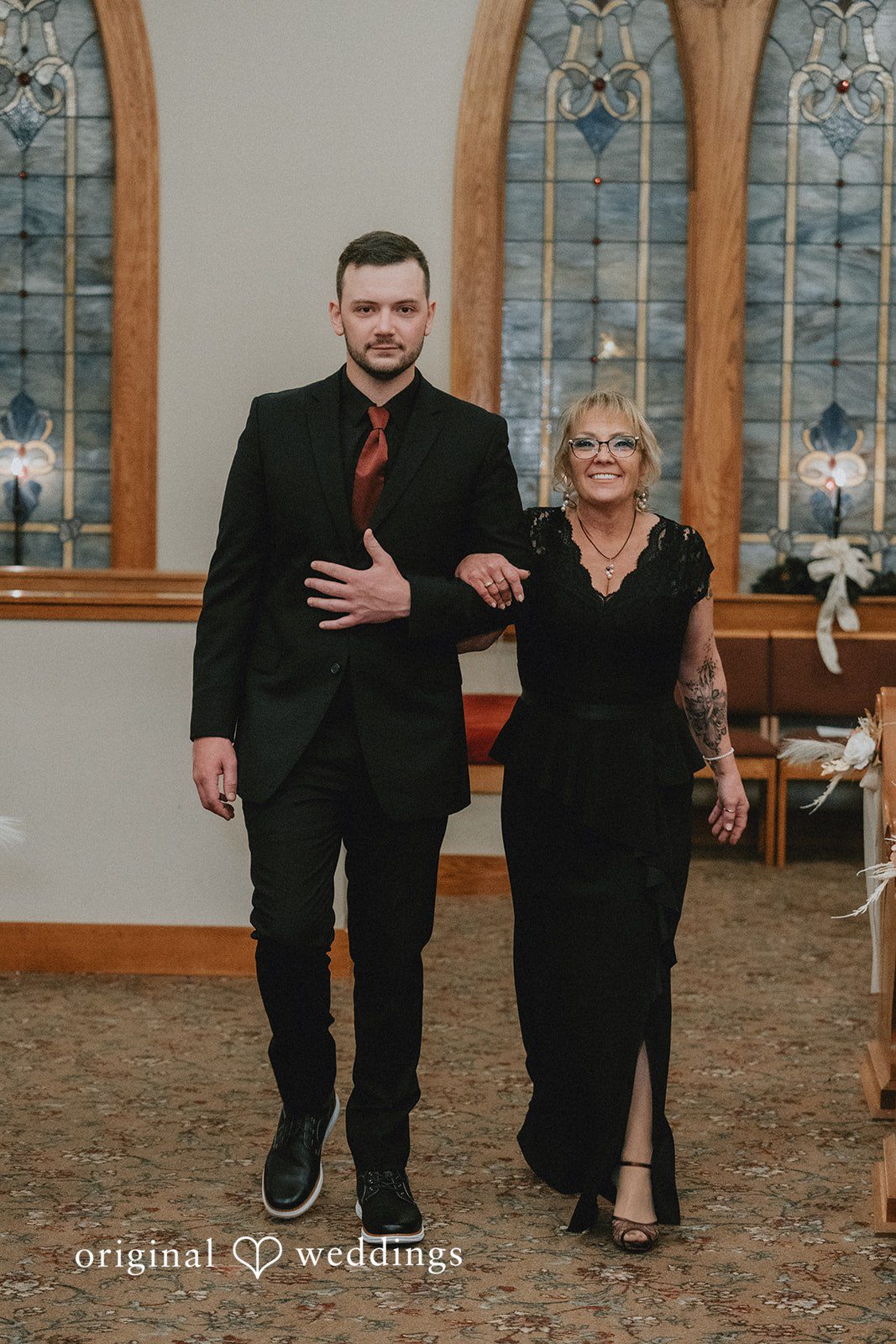 Sarah + Justin A groom in a black suit walks down a church aisle arm-in-arm with a smiling woman in a black dress.