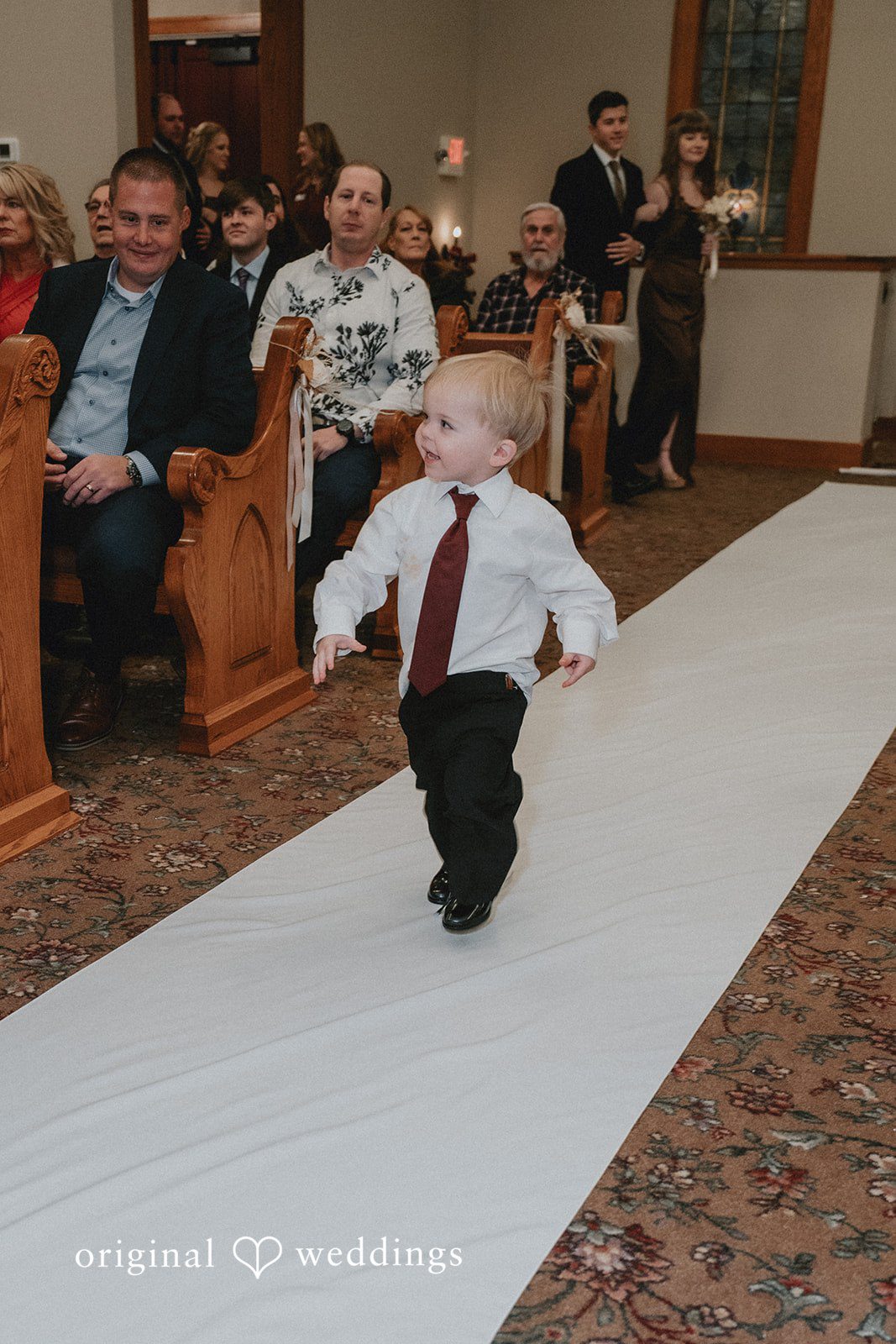 Sarah + Justin A toddler ring bearer wearing a white shirt and red tie smiles while running down a white wedding aisle runner.