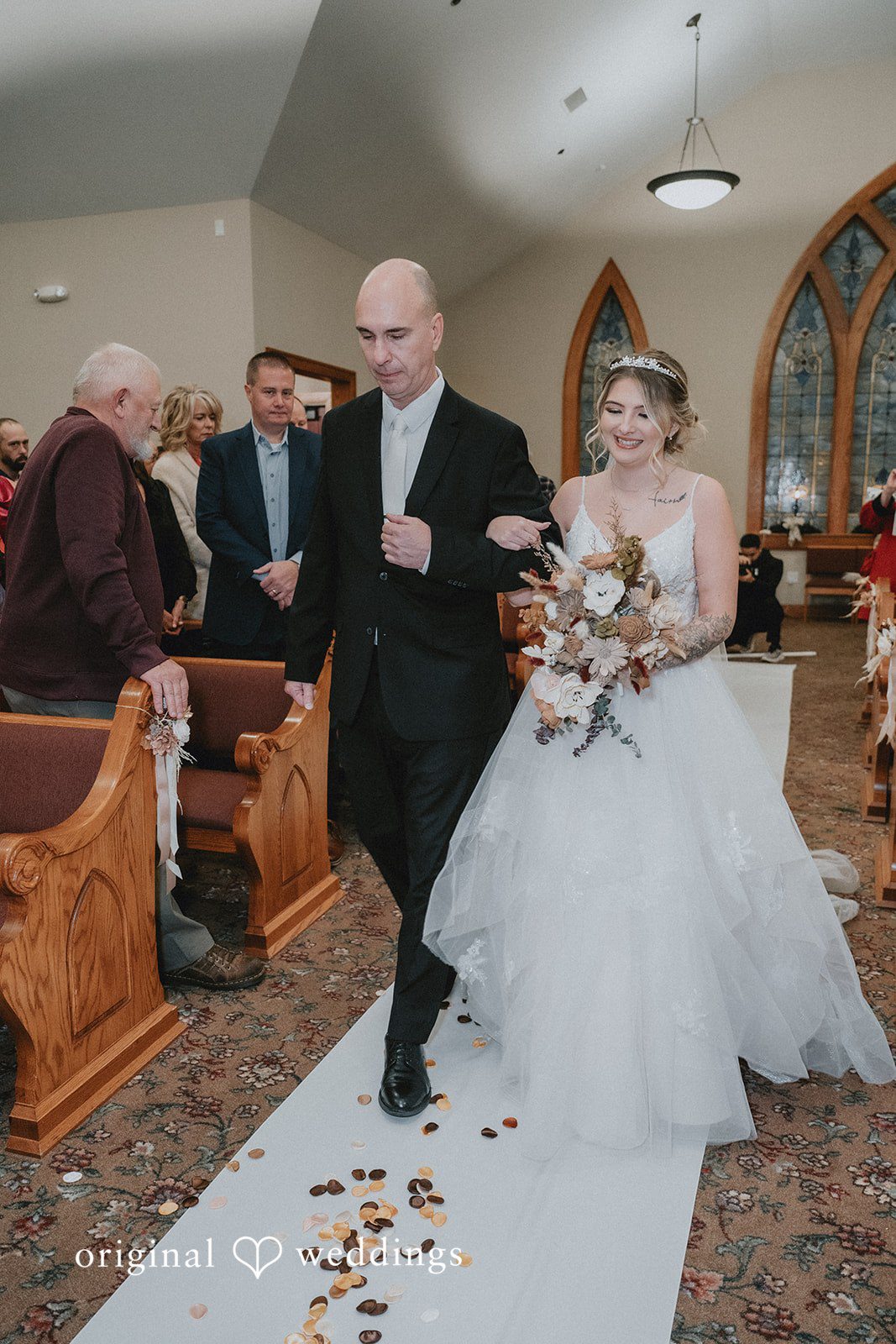 Sarah + Justin A smiling bride walks down a church aisle arm-in-arm with a man in a black suit.