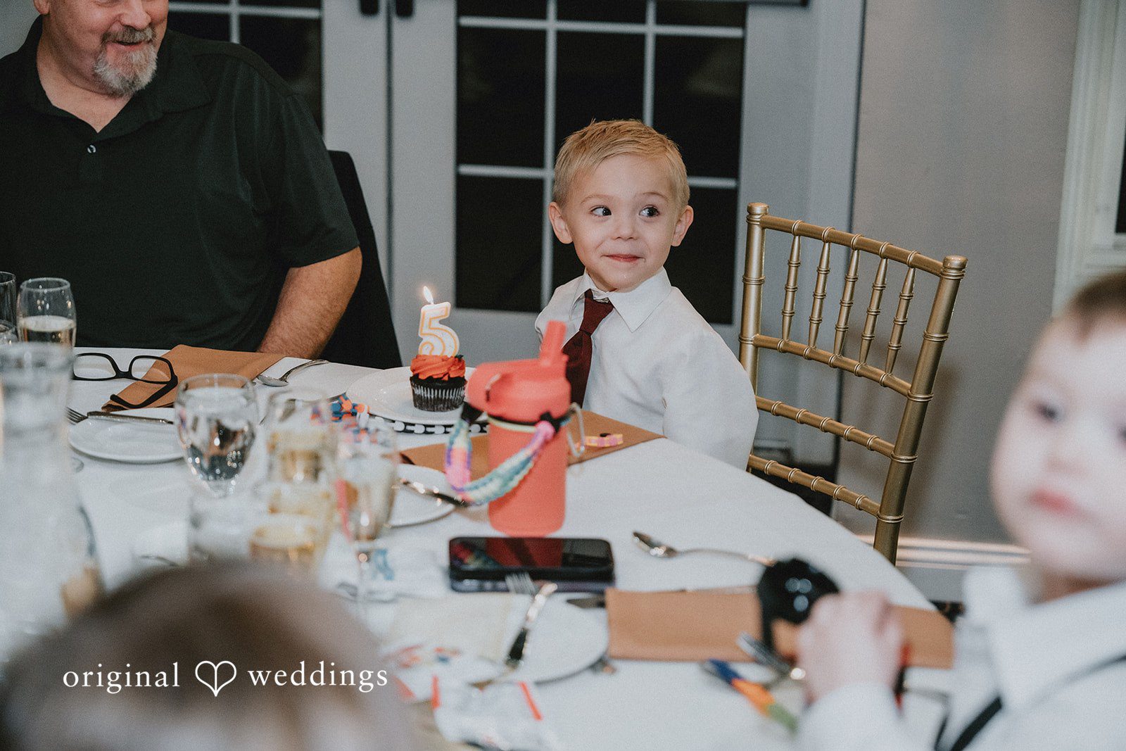 Sarah + Justin A young boy in a white shirt and red tie smiles at a wedding table.