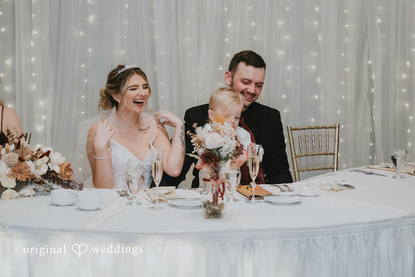 Sarah + Justin A laughing bride and smiling groom sit with a young boy at their wedding head table against a backdrop of glowing fairy lights.