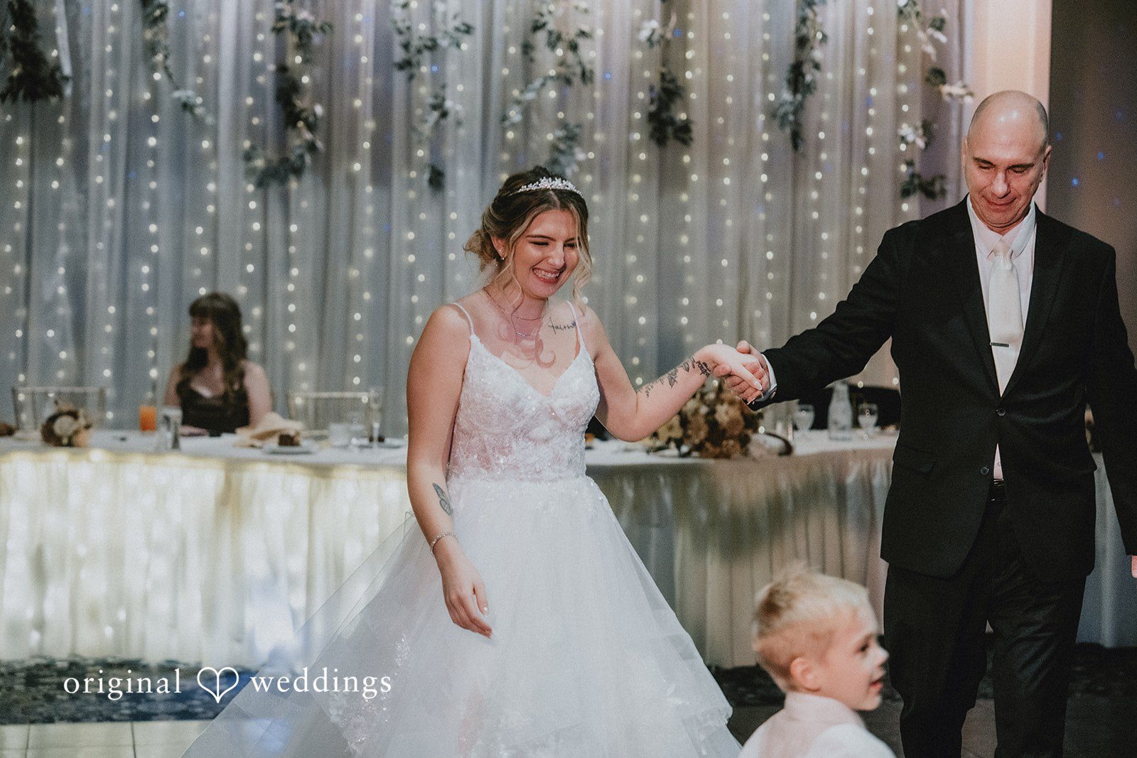 Sarah + Justin A smiling bride holds hands with man in a black suit during their dance in front of a fairy-light backdrop.