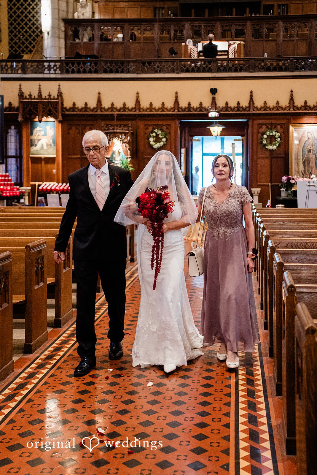 Michael + Maria Bride walking down the church aisle with her parents holding a red bouquet.