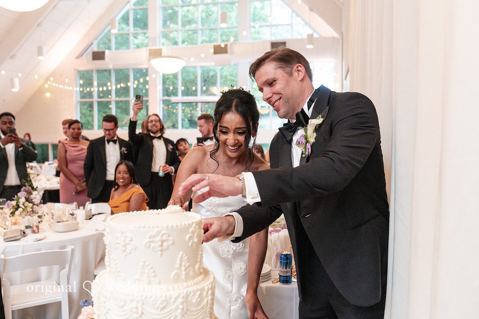 Joyful cake-cutting moment as newlyweds smile surrounded by family and friends.