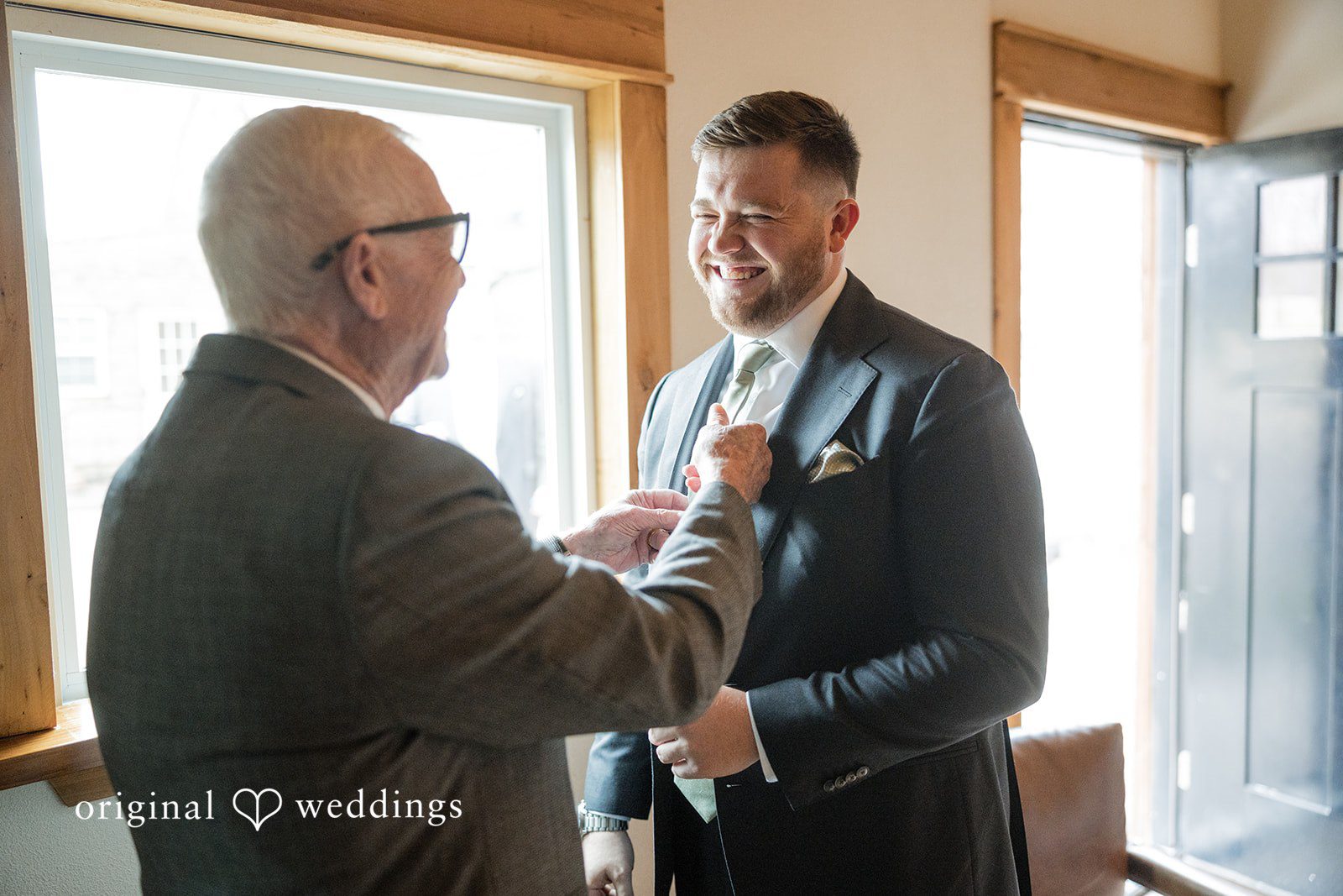 Natalie + Dakin Man adjusting the boutonniere on the lapel of another groom's grey suit jacket.