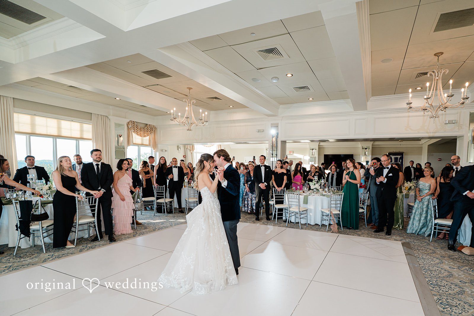 Wide shot of a bride and groom dancing together in the center of a crowded reception hall.