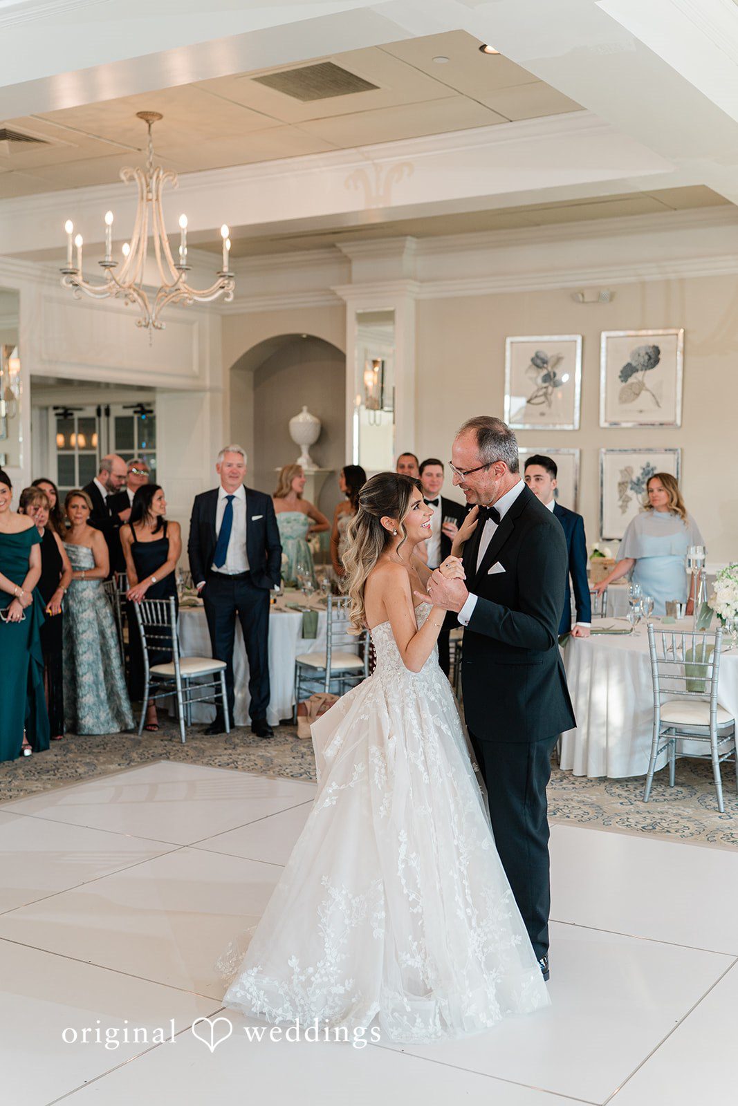 A bride in a white wedding gown and a man in a black tuxedo smile at each other while dancing,