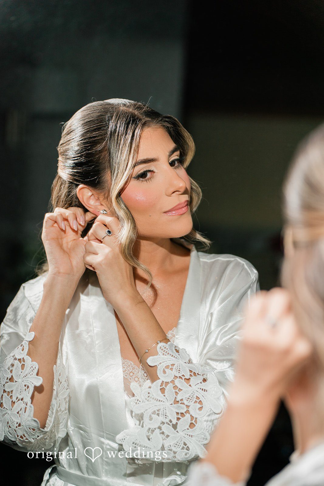 Bridein a white lace robe adjusting her earring while looking into a mirror.