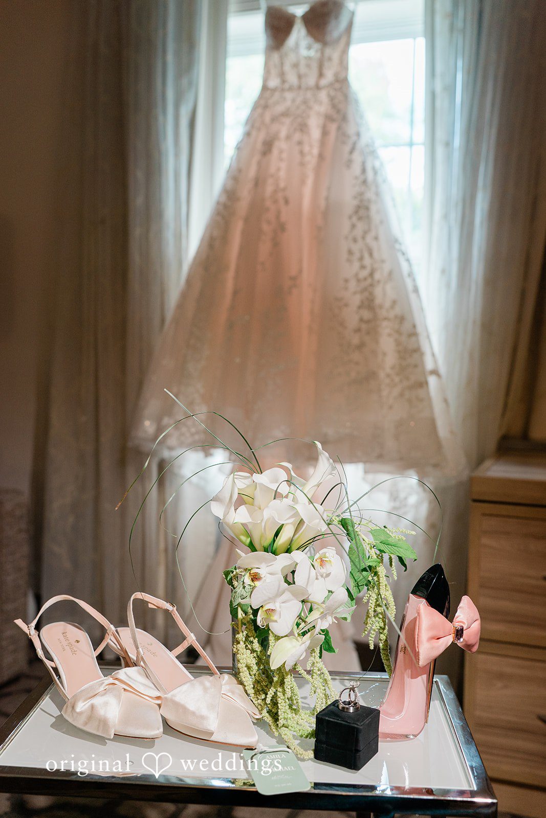 Detailed bridal setup showing a hanging wedding dress, white shoes, and floral arrangements on a table.