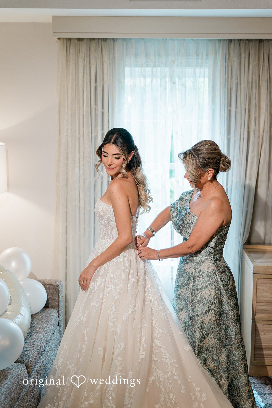 Woman in a patterned dress helping a bride fasten the back of her white wedding gown.