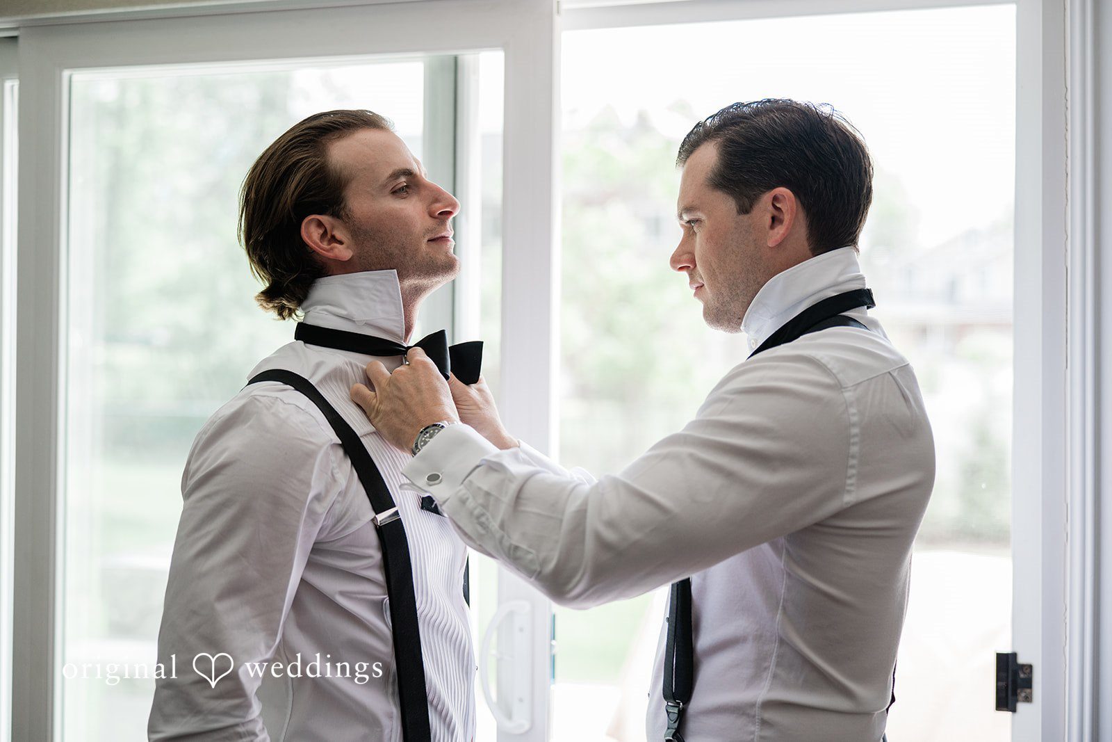 Man in a white shirt helping a groom adjust his black bowtie.