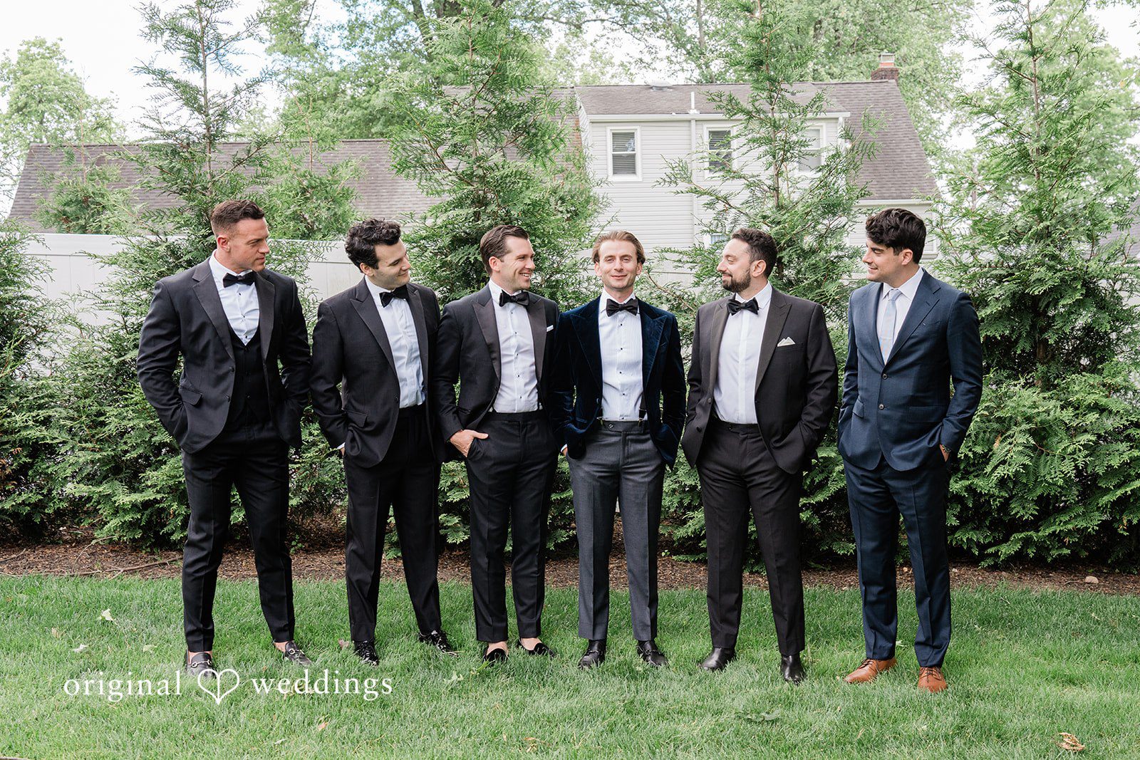 Groom and groomsmen dressed in black tuxedos standing in a line on a grassy lawn.