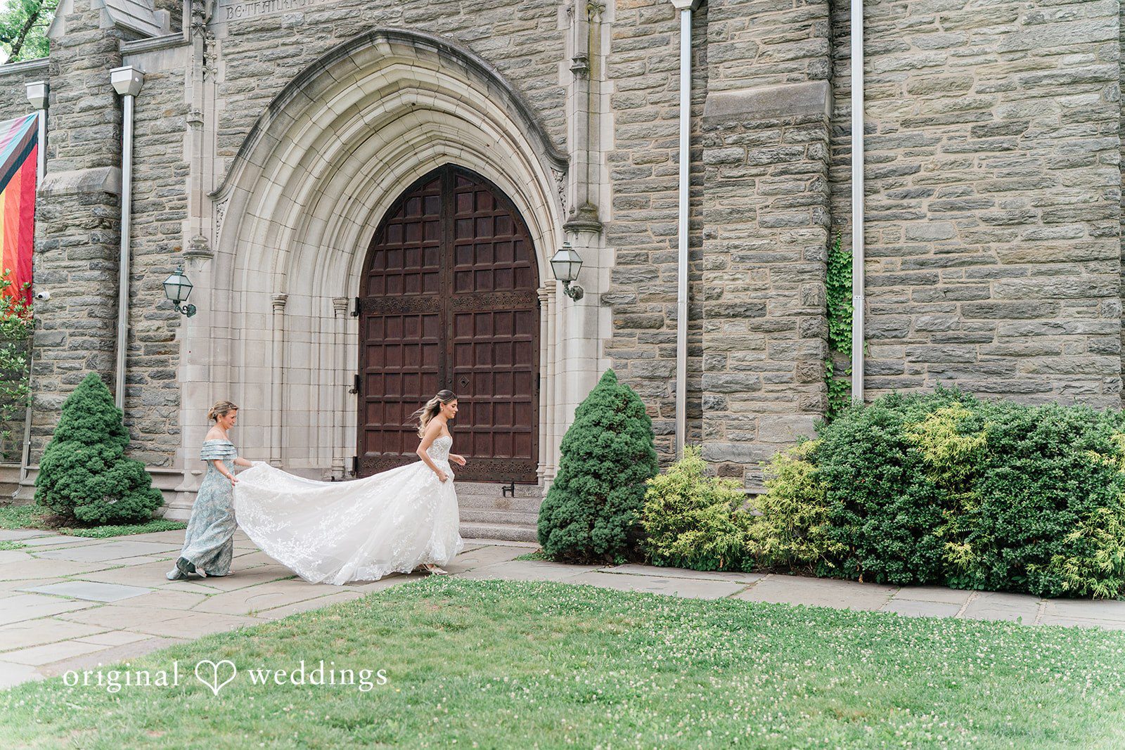 A woman carries the train of a bride's gown past a stone building with a large arched wooden doorway.
