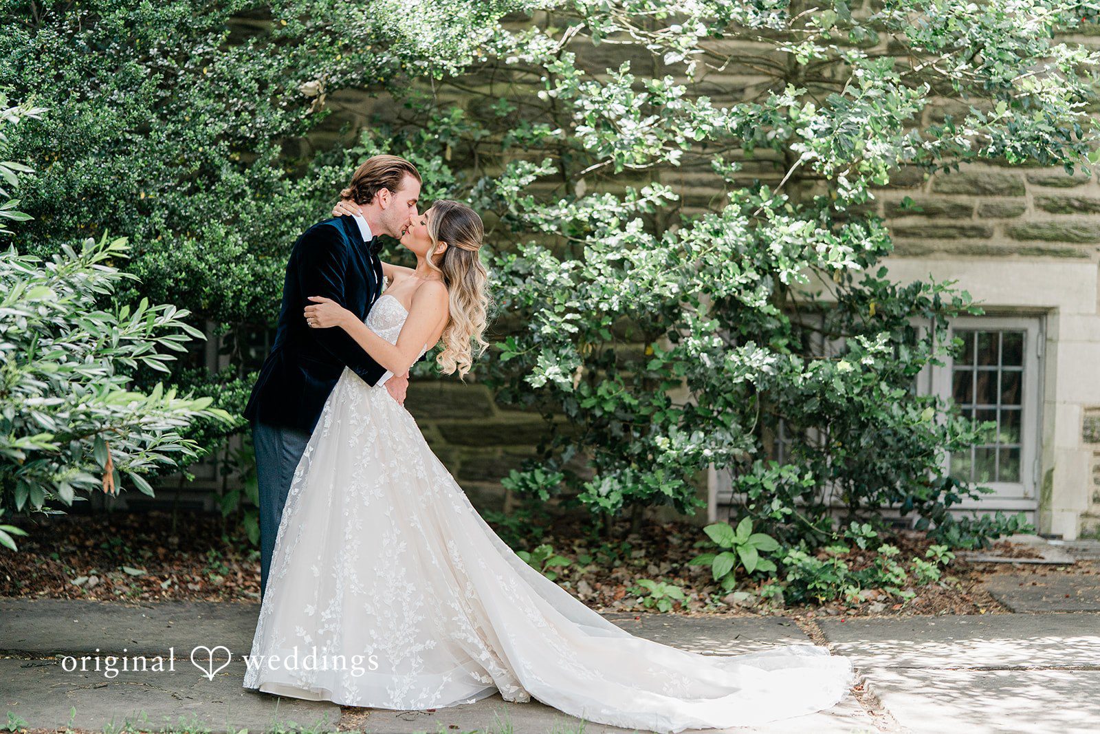Groom kissing the bride on a pathway next to green bushes.