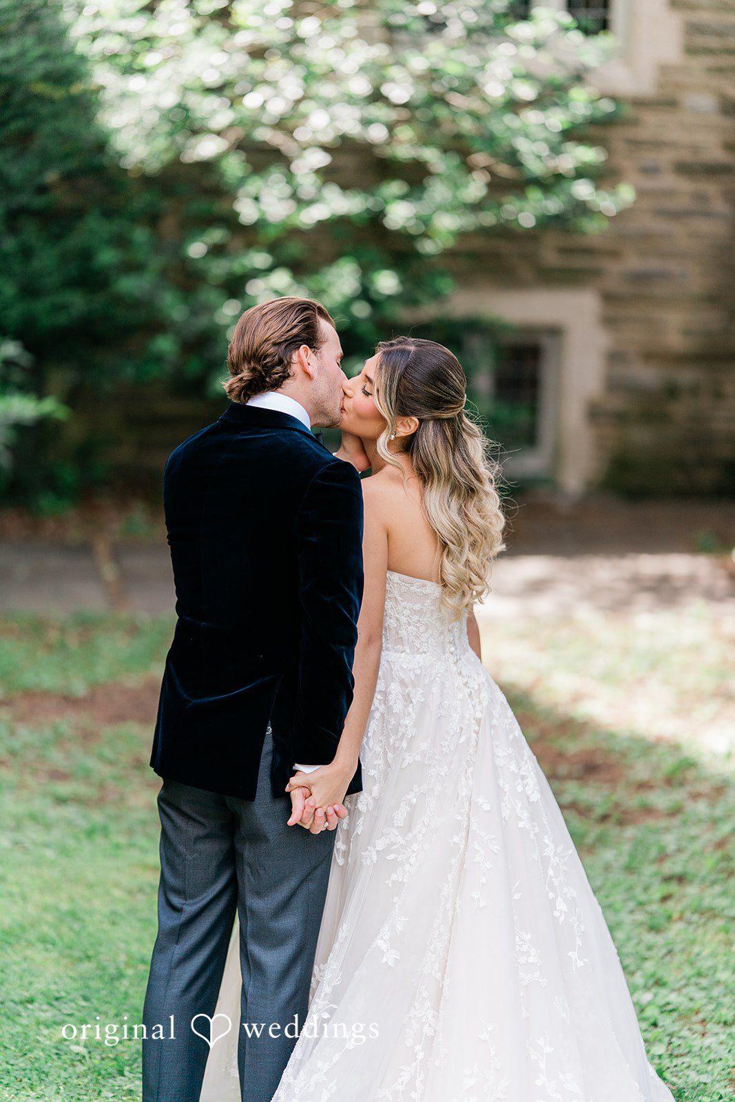 Groom kissing the bride while standing on a grassy path near a stone building.