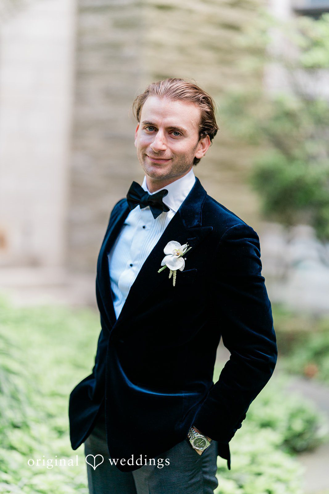 Groom in a black velvet tuxedo jacket posing with a smile against a soft green background.