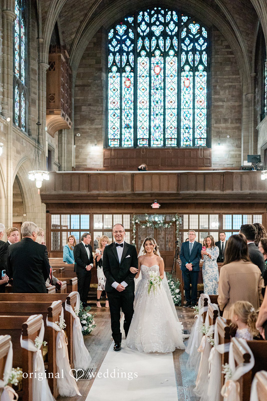 Bride walking down a church aisle flanked by wedding guests sitting in wooden pews.