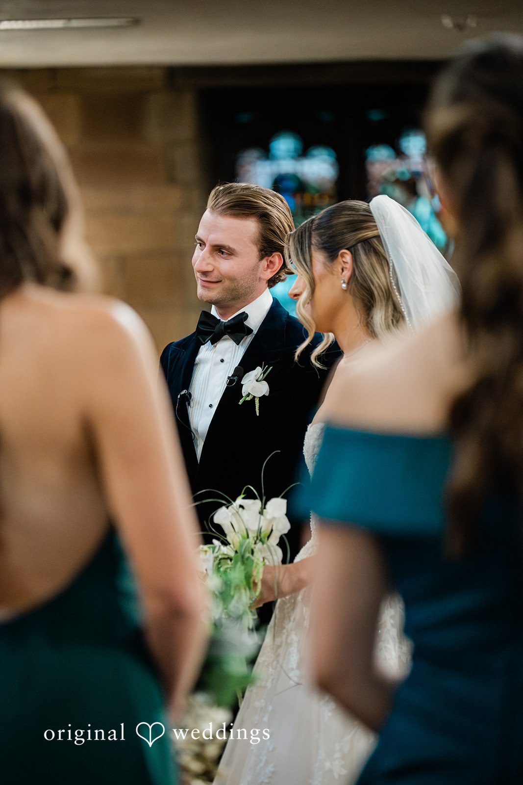 A groom in a black tuxedo looks smilingly during their wedding ceremony