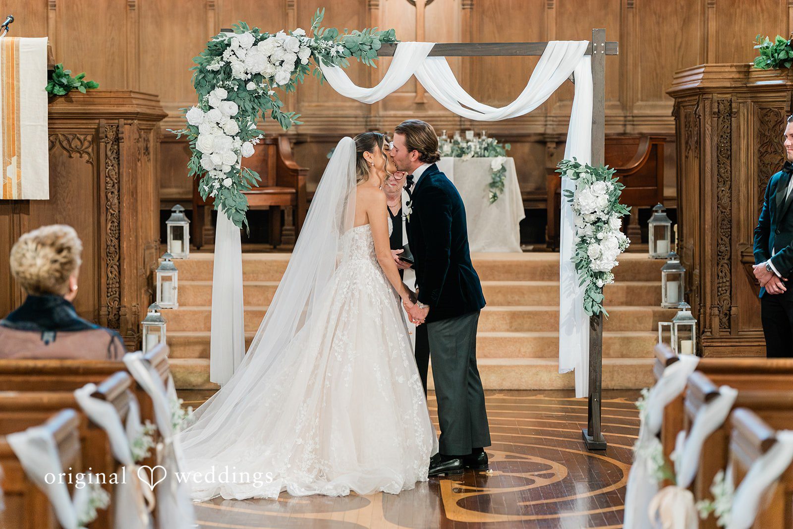 Bride and groom sharing their first kiss at a decorated white wedding altar.