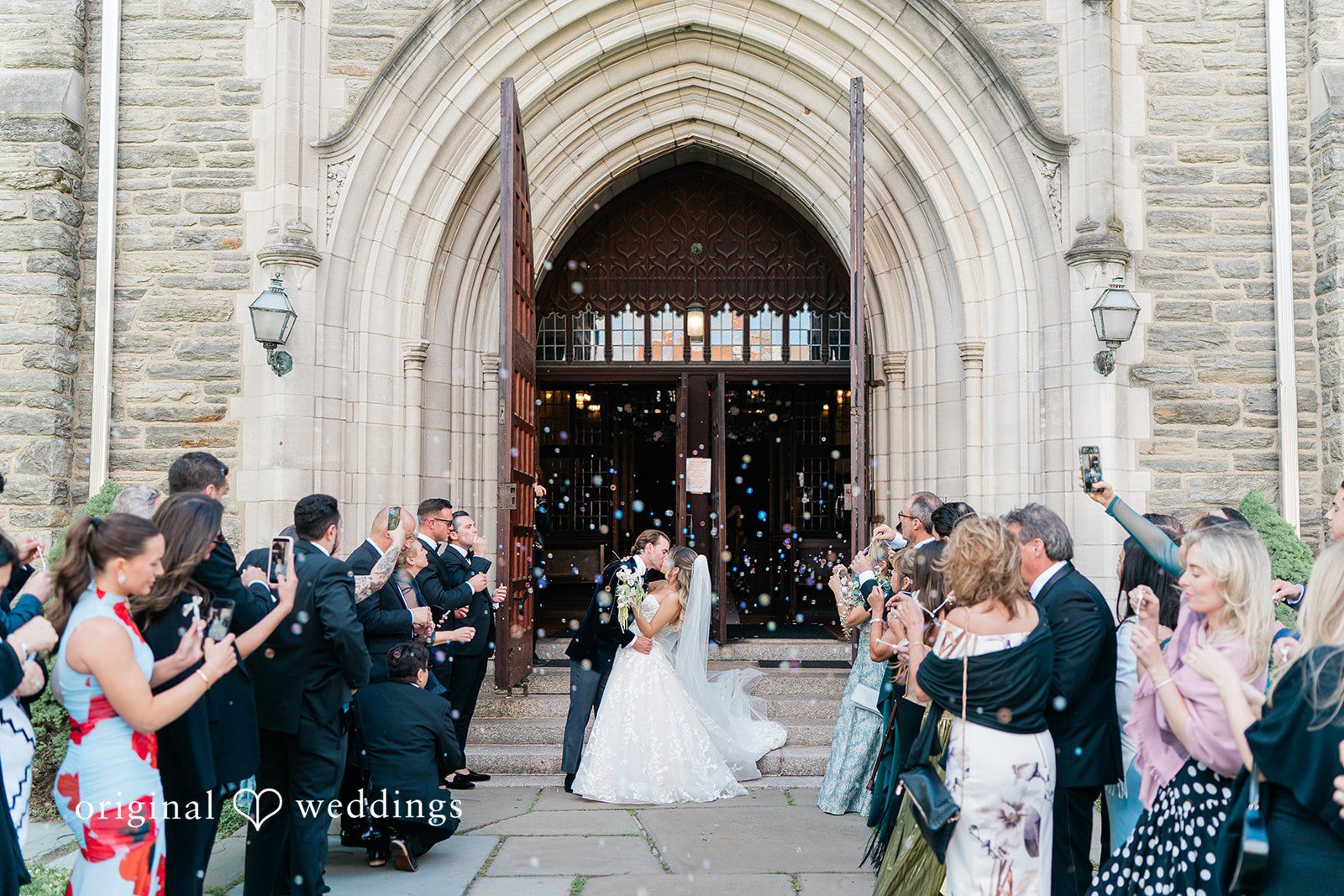 Newlyweds walking out of a grand stone church building into a crowd of cheering guests.