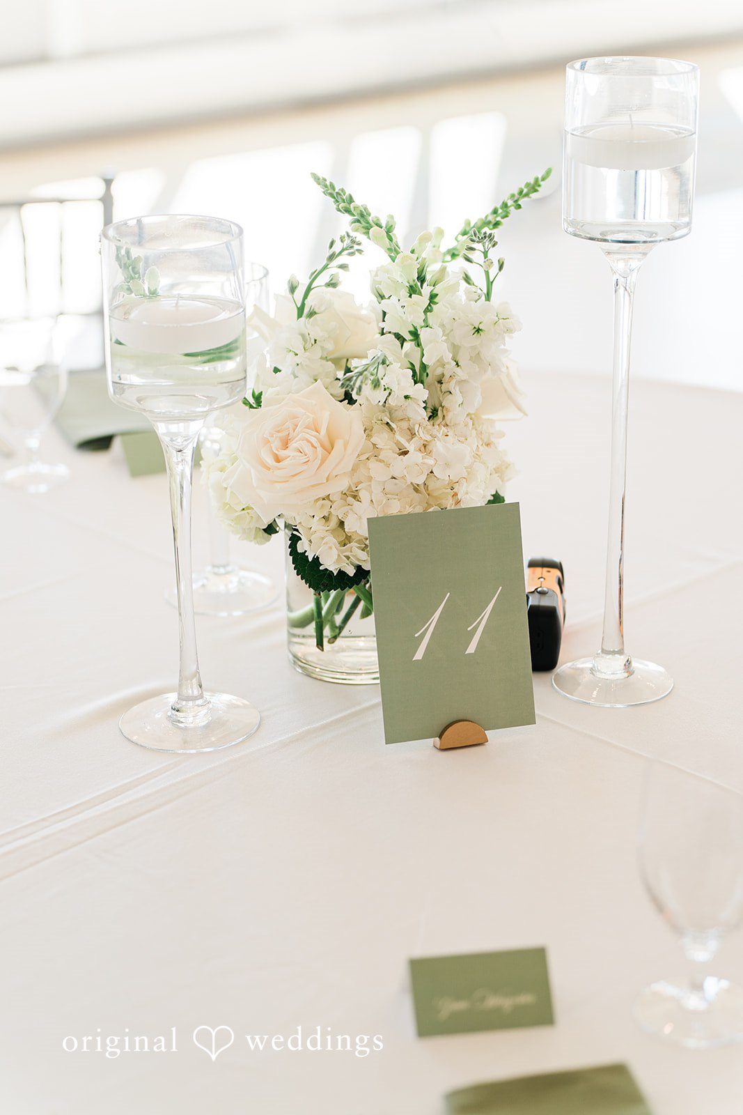 Minimalist reception table setting featuring white floral arrangements, tall wine glasses, and olive-green place cards.