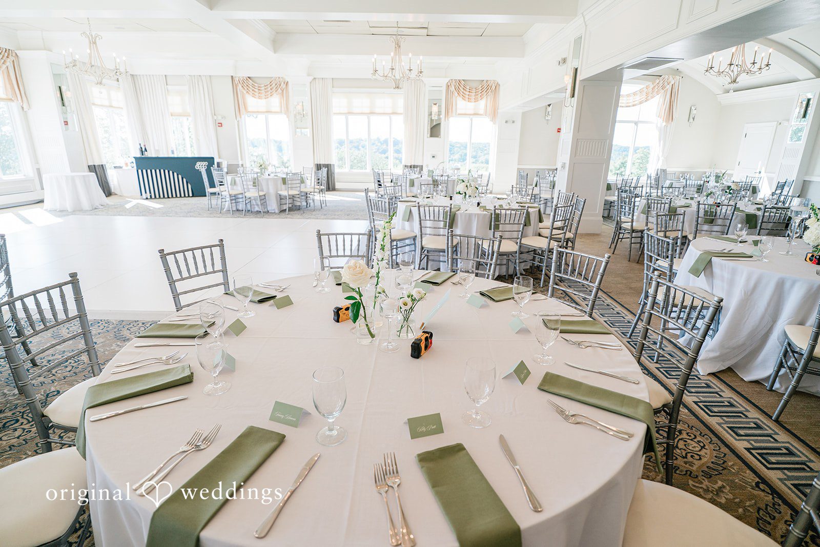 Round reception tables set with white linens, green napkins, and clear acrylic chairs.